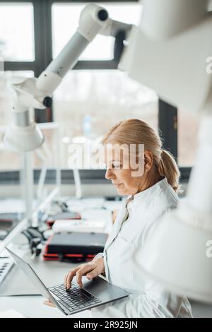 Scientist using laptop by robotic arms in laboratory Stock Photo