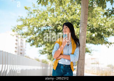 Thoughtful businesswoman leaning on tree holding smart phone Stock Photo
