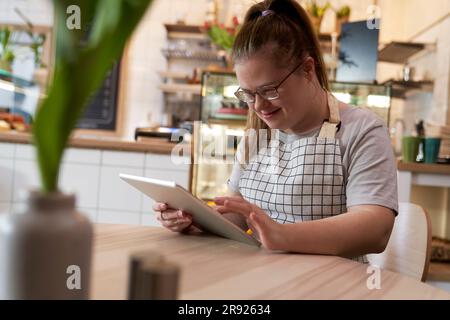 Smiling cafe owner with down syndrome using tablet PC at table in coffee shop Stock Photo