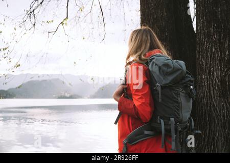 Woman with backpack standing next to tree near lake Stock Photo