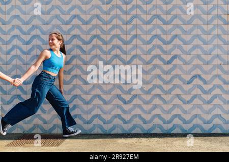 Happy girl holding hand of sister and running in front of wall Stock Photo