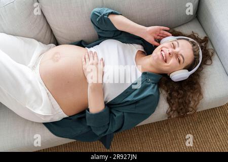 Happy pregnant woman listening to music lying down on sofa at home Stock Photo