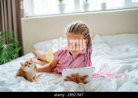 Girl holding tablet PC and petting ginger kitten on bed Stock Photo