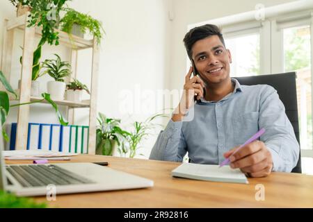 Smiling young businessman talking on smart phone and writing notes in office Stock Photo