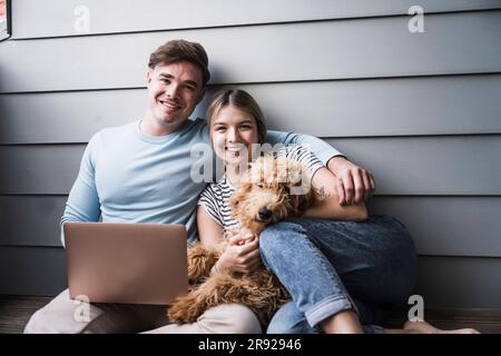 Happy young couple sitting with dog in front of wall Stock Photo