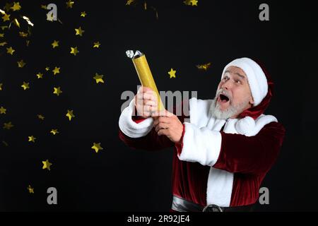 Emotional man in Santa Claus costume blowing up party popper on black background Stock Photo