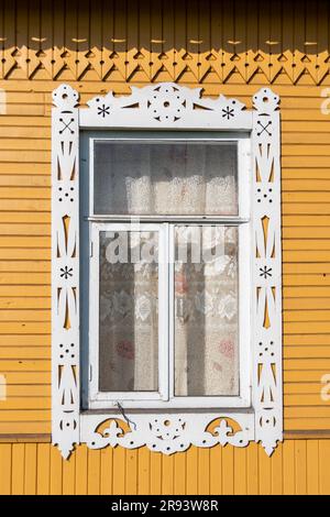 Close-up of a window framed with a carved wooden jamb decorated with traditional white patterns on the facade of a yellow painted Russian isba Stock Photo