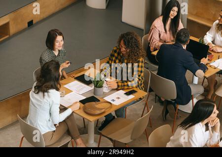 Three colleagues, male and females, having fun conversation and smiling while working at the cafe of modern co-working space Stock Photo