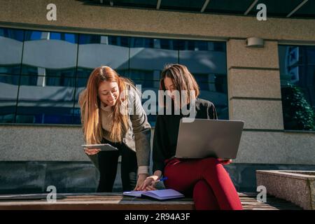 Front view shot of two businesswomen working on their lap top and tablet. They are writing notes down in a notebook Stock Photo