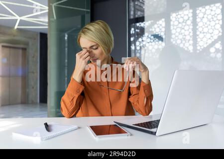 Overtired and overworked woman at workplace inside office, businesswoman took off glasses has severe pain in head and eyes, female worker works long time at laptop. Stock Photo