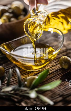 Pouring olive oil from carafe to the glass bowl and in background are olive branches put on table. Stock Photo