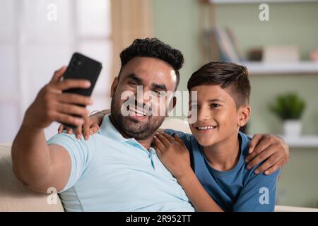 Happy smiling indian father with son taking selfie on mobile phone on sofa at home - concept of social media sharing, cyberspace and family time. Stock Photo