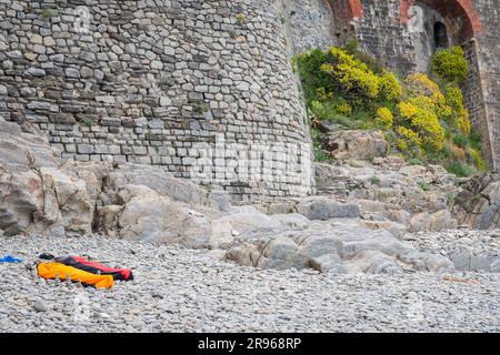 Manarola Italy - April 25 2011; Manarola ancient fishing village stony beach with two people sleeping in bags by Mediterranean sea at base of large st Stock Photo