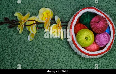 Yarn for crocheting. Top view, web banner with copy space. Several woolen multicolored balls of thread in wicker basket on green crocheted background. Stock Photo