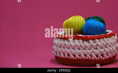 Concept craft handmade needlework hobby from home. Yarn for crocheting. Several woolen multi colored balls of thread in a wicker basket on a pink back Stock Photo