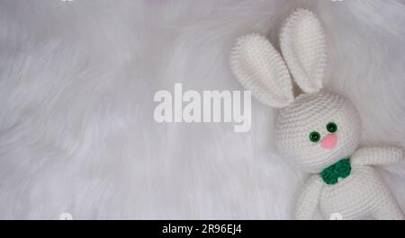 Crochet stuffed animals. Charming white knitted rabbit with green bow tie and a pink nose lies on a fluffy wool background. Top view, web banner with Stock Photo