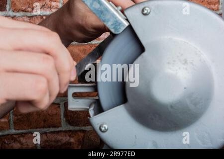 A knife sharpener and hand with a blade on a wooden table, close-up against a brick wall background Stock Photo