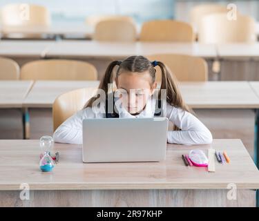 Caucasian girl sits at a desk at school and studies at a laptop. Hourglass on the table. Stock Photo