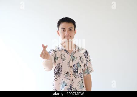 A young Asian man wearing a pink shirt with tropical patterns is posing as 'Saranghae' usually known as finger heart. Focused on the man. Stock Photo