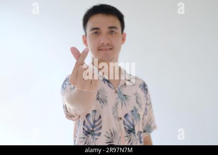 A young Asian man wearing a pink shirt with tropical patterns is posing as 'Saranghae' usually known as finger heart. Focused on the finger. Stock Photo