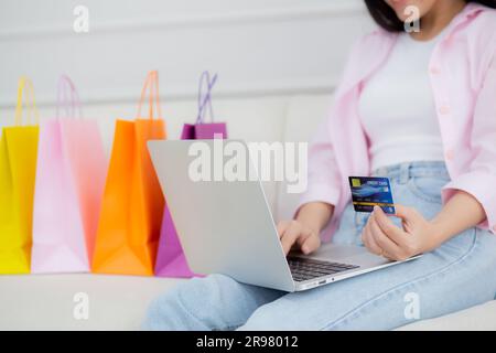 Closeup hands of woman sitting on sofa using laptop computer shopping online with credit card while paper bag on sofa, female paying with transaction Stock Photo