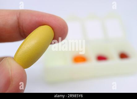 Fingers Holding a Yellow Supplement Pill with Blurry Pill Organizer Case in the Backdrop Stock Photo