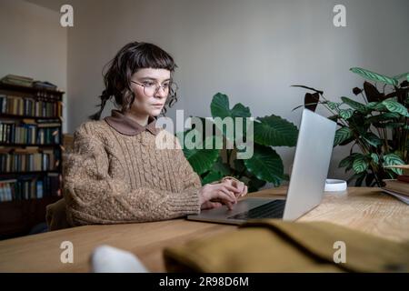 Focused teen student girl wearing glasses sitting at table typing on laptop Stock Photo