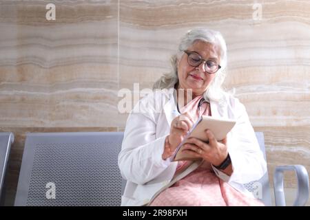 Portrait of smart senior female doctor in white lab coat sitting on bench  with a note pad in hand. writing pad. taking notes. Stock Photo