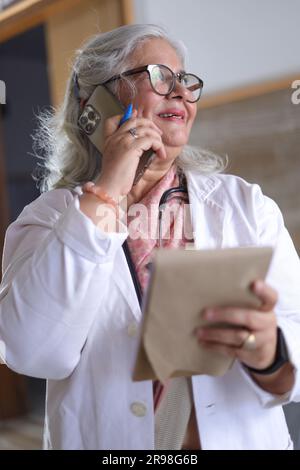 Portrait of smart senior female doctor in white lab coat talking over a mobile phone call with a note pad in hand. writing pad. Stock Photo