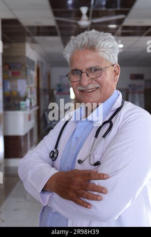 Portrait of smiling male doctor in lab coat with arms crossed. Happy senior doctor in white coat smiling on hospital corridor background. Stock Photo