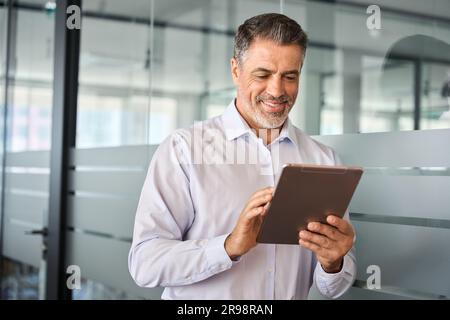 Smiling busy mature male manager using tablet standing in office. Stock Photo
