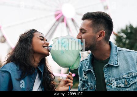 Young guys are having fun in an amusement park. Girlfriend and her boyfriend biting cotton candy during the festival. Stock Photo