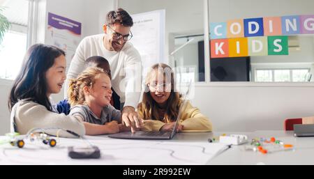 Male teacher guides elementary school students in a robot programming class. Kids learn to code using education software on a laptop and control robot Stock Photo