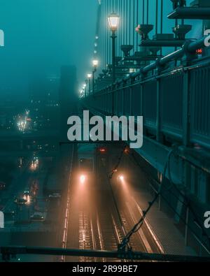 Three illuminated traffic lights hang on a bridge over a foggy city street at night, with a view of the illuminated downtown buildings in the distance Stock Photo