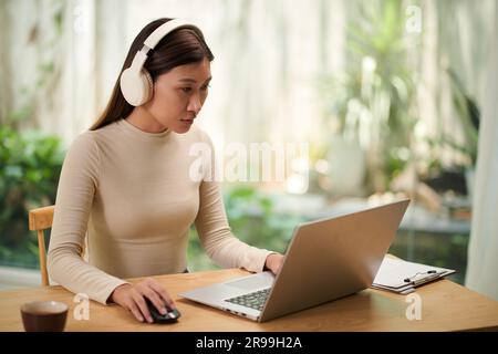 Young woman wearing headphones when working on laptop and aswering e-mail from coworkers Stock Photo