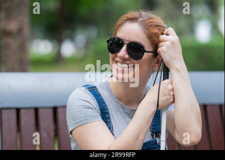 Blind red-haired woman sitting outdoors leaning on a cane.  Stock Photo