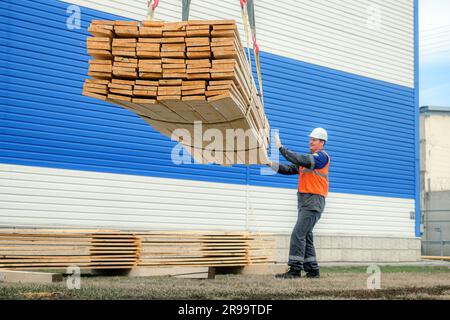 A slinger unloads wooden planks into a pile. A worker in a helmet and a high-visibility vest unloads new boards outside in the open air. Sale of lumber for construction. Authentic workflow. Industrial background. Stock Photo