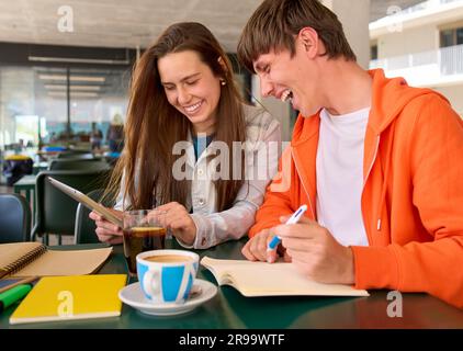 Young Caucasian female and male laughing while using tablet notepad in university campus cafeteria.  Stock Photo