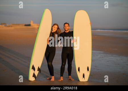 Happy Young Couple Posing With Their Surfboards On The Beach At Sunset Stock Photo