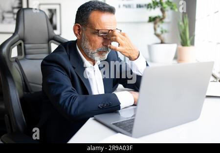 Unhappy sad tired mature european man in suit work on laptop, has stress, migraine in office interior Stock Photo