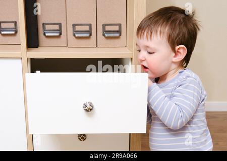 Toddler baby opened the cabinet drawer. Child boy play with wardrobe in the home living room. Kid age one year nine months Stock Photo