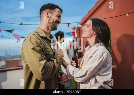Smiling young couple flirting and having fun at a rooftop party with friends, drinking beer, enjoying and having a pleasant conversation. Handsome guy greeting his joyful girlfriend at celebration. High quality photo Stock Photo