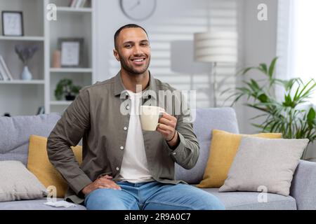 Portrait of a young hispanic man sitting on the sofa at home and holding a cup with a drink. He looks at the camera with a smile. Stock Photo