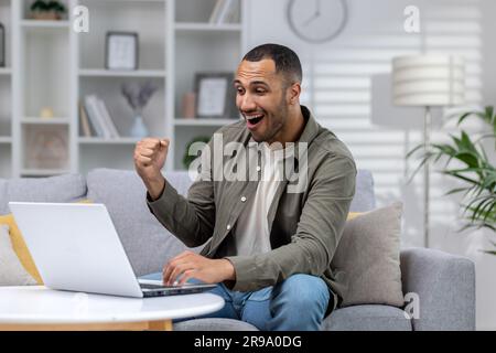 Happy young african american man using laptop on couch at home. Celebrates, rejoices, received good news, watches online sports games. Stock Photo