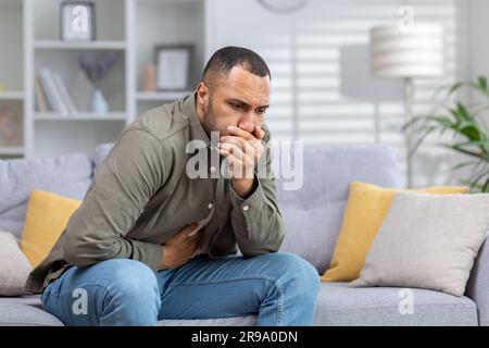 A young African American man is not feeling well. Sits at home on the couch, holds his stomach with his hand, covers his mouth with his hand, wants to vomit, feels nausea and severe pain. Stock Photo