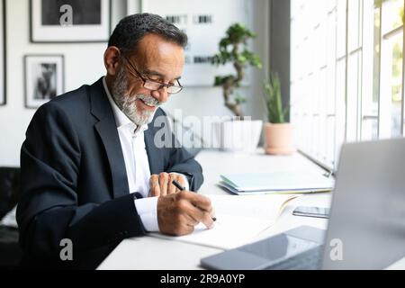 Cheerful mature european man in suit work on laptop, making notes, signing contract in modern office Stock Photo