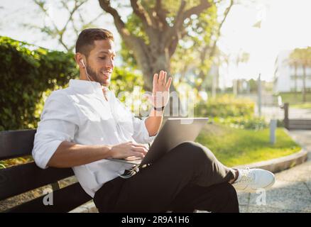 Excited businessman in formal wear looking at laptop webcam and greeting client, sitting outdoors on bench, copy space Stock Photo