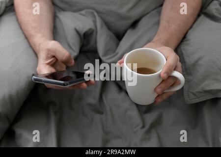 hands of man with cell phone and cup of coffee in bed Stock Photo