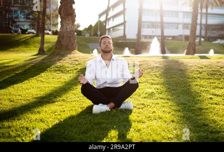 Calm young businessman in formal wear meditating with closed eyes outdoors, practicing yoga, sitting on lawn in park Stock Photo