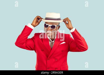 Funny smiling dark skinned man in stylish red jacket adjusts his hat on pastel light blue background Stock Photo